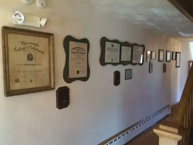Wall lined with framed certificates and plaques in a narrow hallway with a wooden staircase on the right