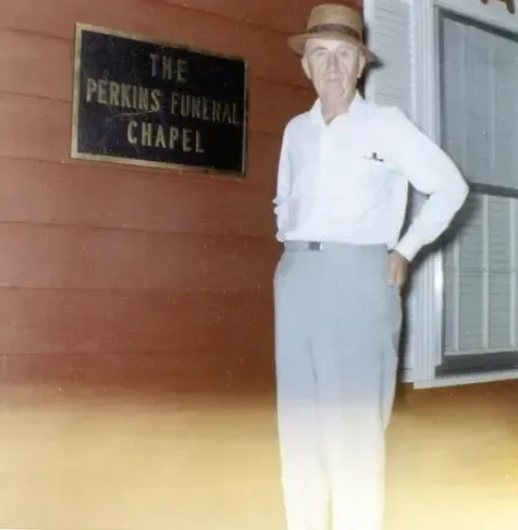 Man standing by a sign reading “The Perkins Funeral Chapel” outside a building.