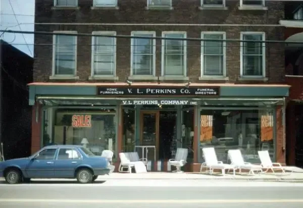 Storefront with a blue car parked outside and white chairs on the sidewalk under an awning.