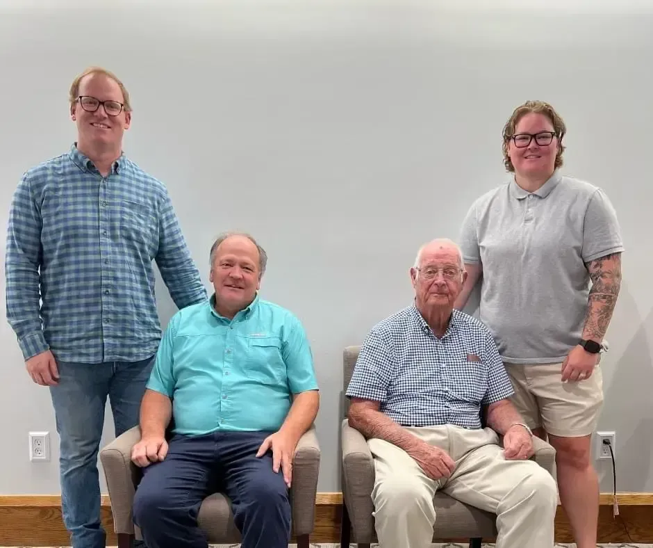 Four adults posing indoors against a gray wall, two seated in front and two standing behind them.