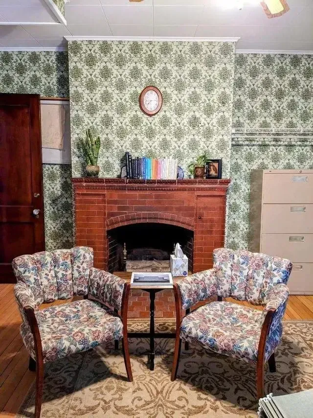 Cozy living room with two floral armchairs, a brick fireplace, patterned wallpaper, and a small table with books.
