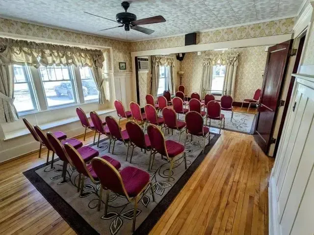 Conference room with rows of red chairs, hardwood floor, ceiling fan, and large windows with curtains