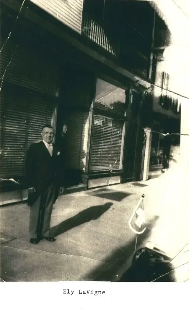 Man standing on a city sidewalk beside storefronts, with a small dog nearby in bright sunlight.