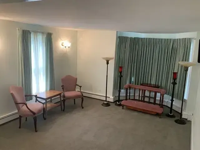 Empty living room with two pink chairs, small table, floor lamps, and a curtained window.