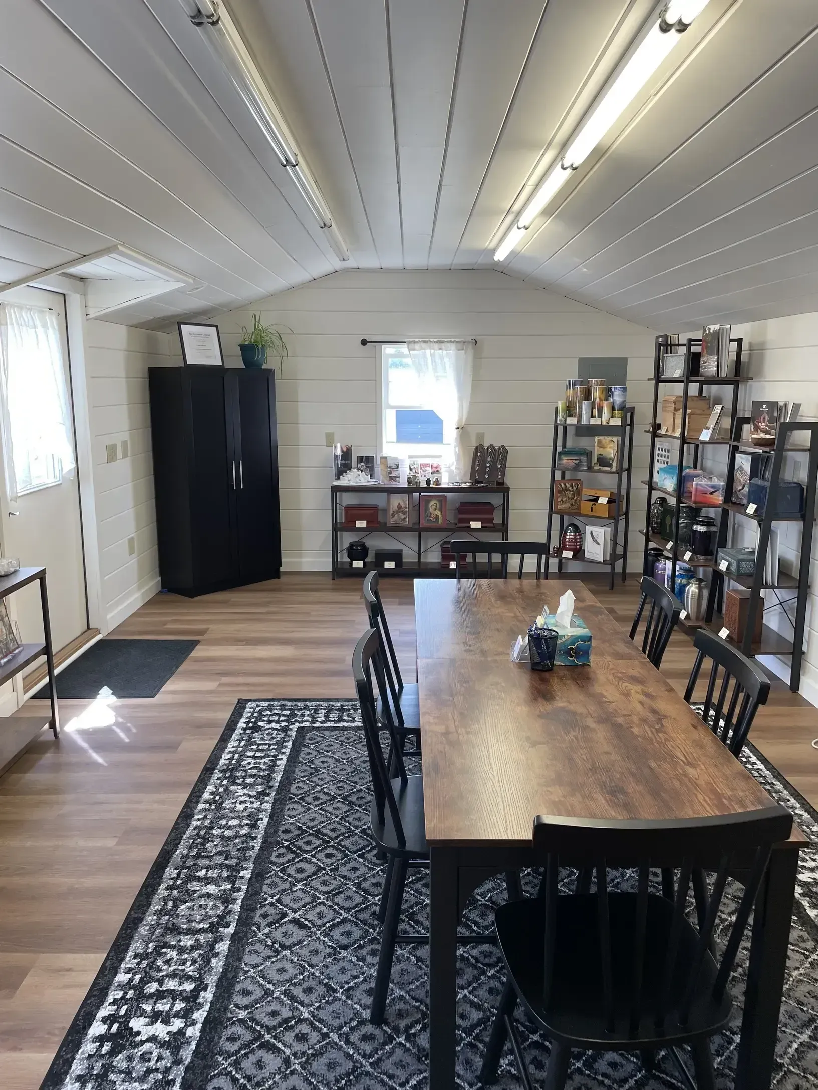 Dining room with wooden table and chairs, patterned rug, shelves, and storage cabinets in a bright white room.