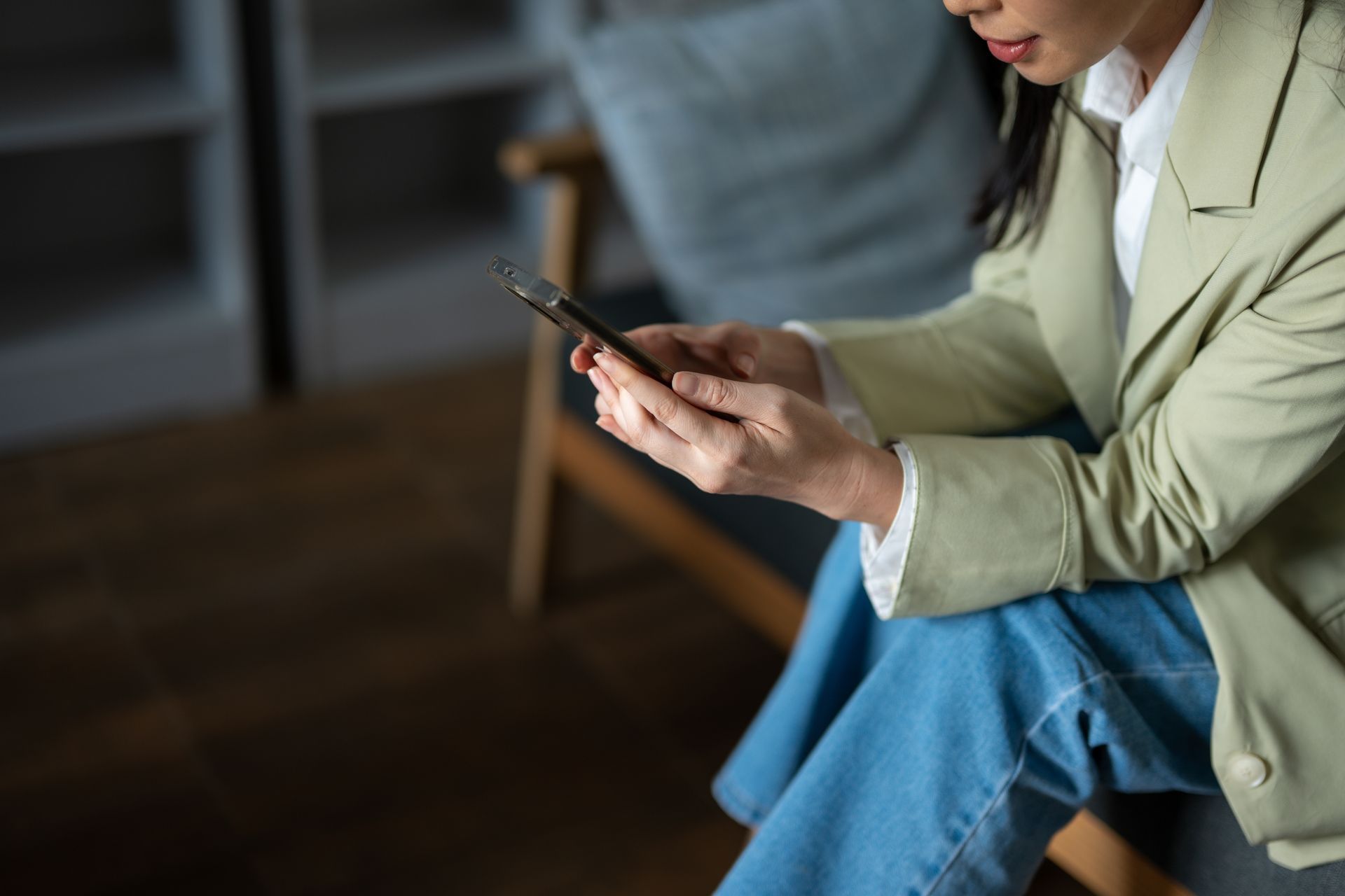 Person in a beige jacket and blue jeans looking at a smartphone while seated indoors