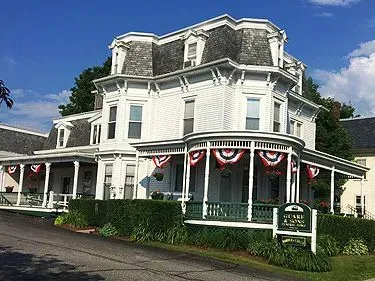 Victorian-style white house with porch, red-white bunting, and a sign in front under a blue sky