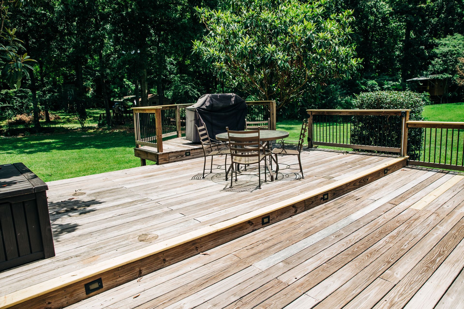Wooden deck with outdoor table, chairs, grill, and railing overlooking a green yard with trees