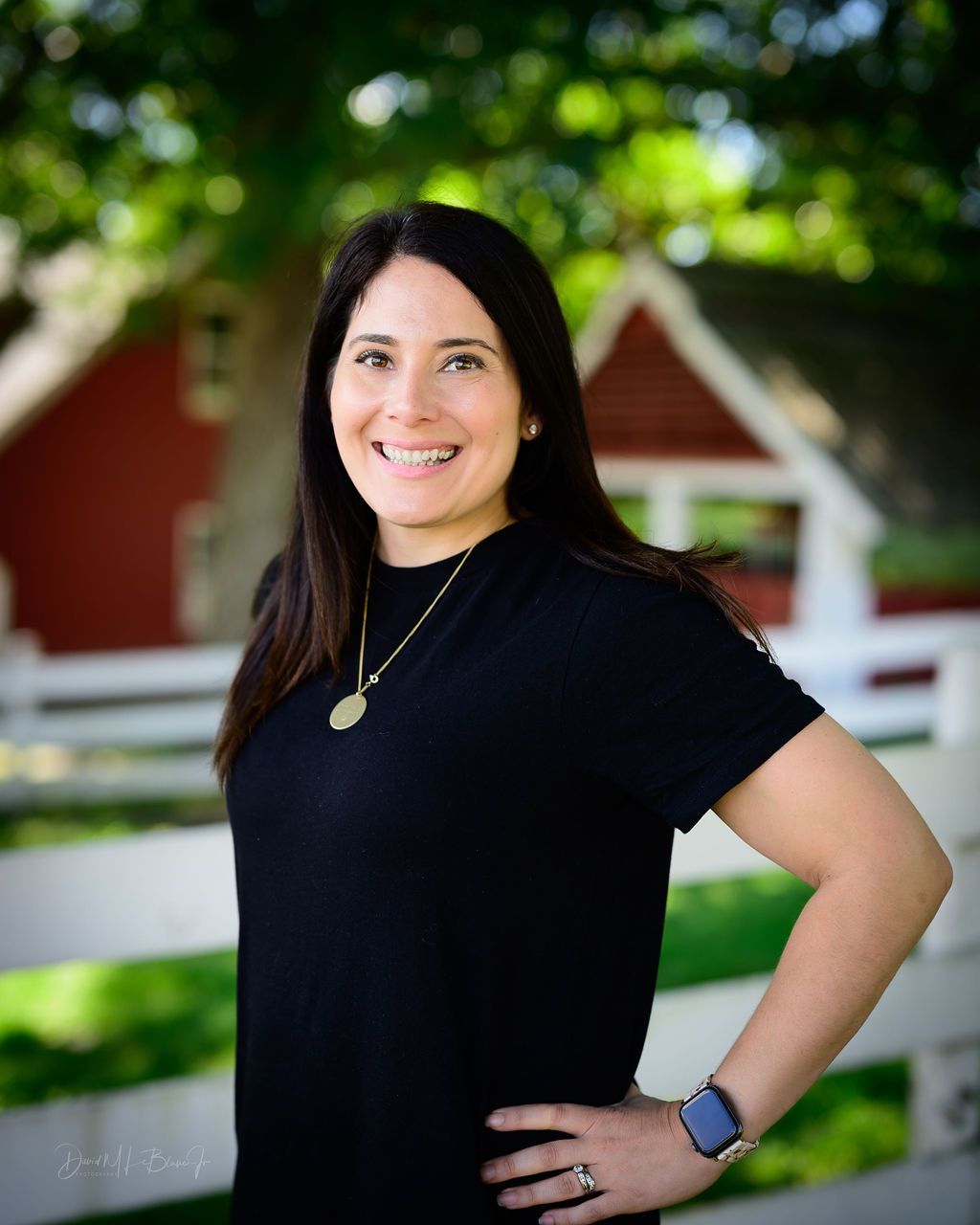 Woman with dark hair smiles in front of a red building and white fence. She wears a black shirt and a watch.