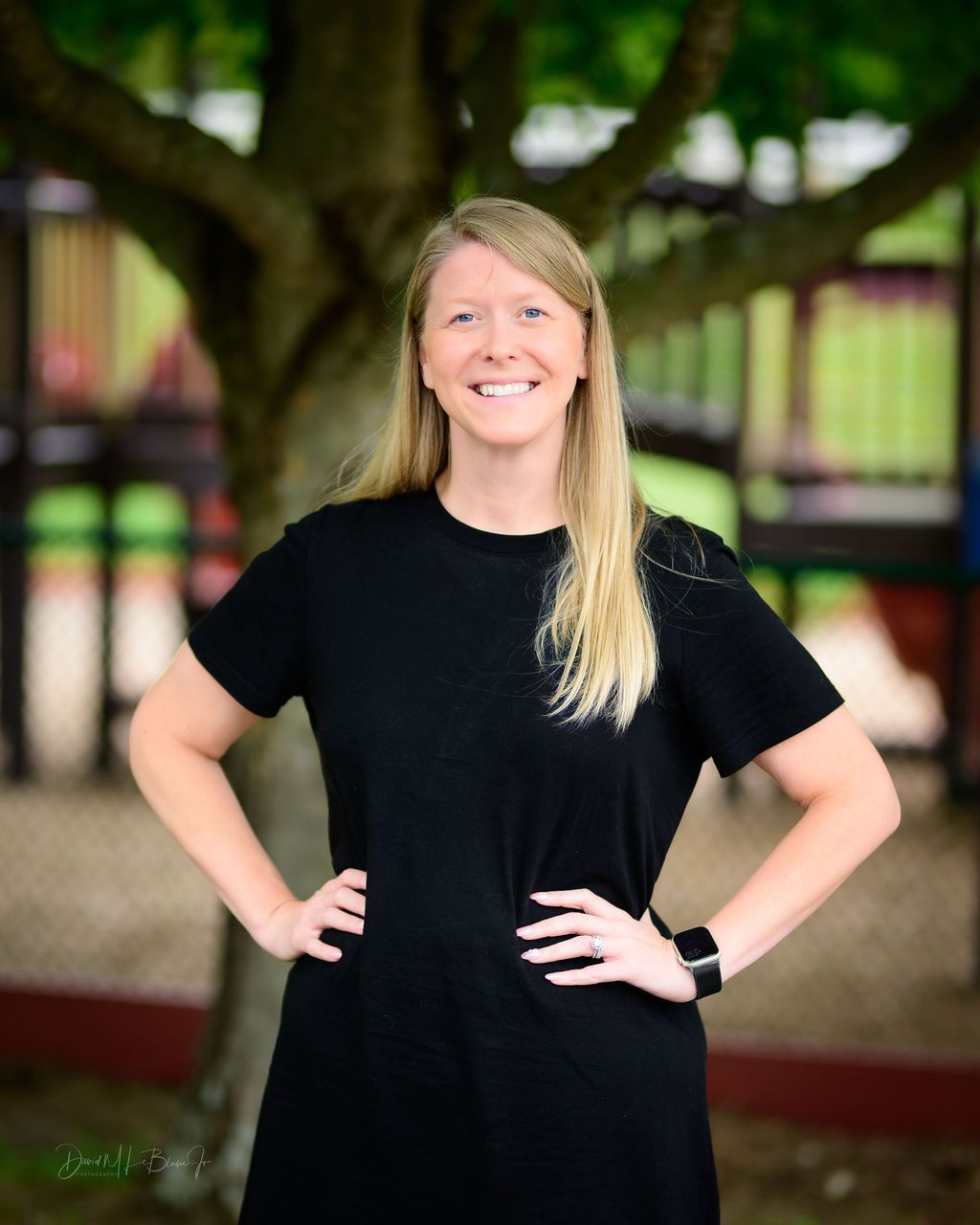 Woman with blonde hair smiling, hands on hips, wearing black dress, in front of a playground.