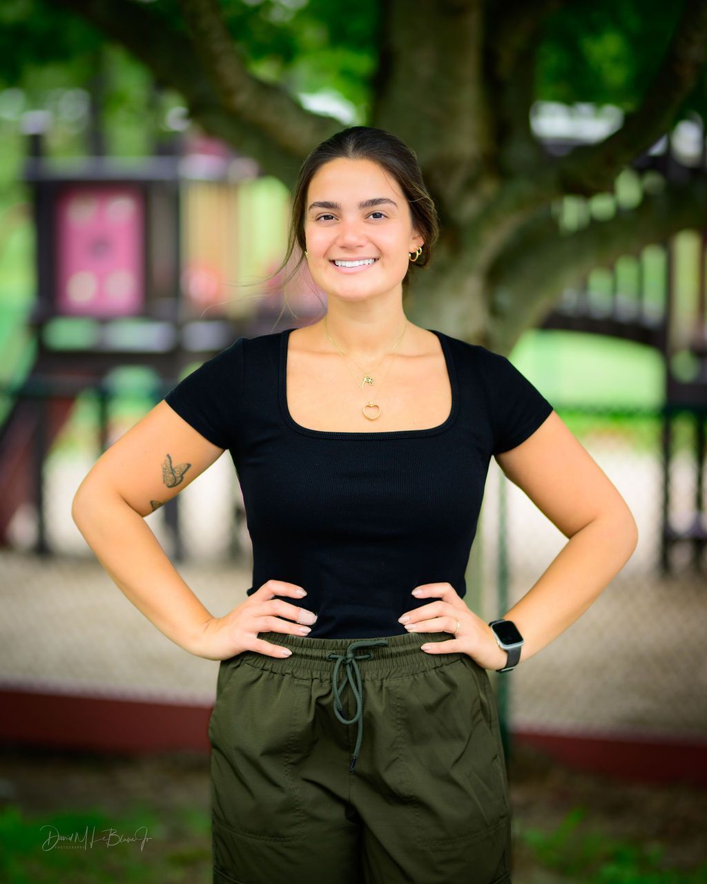 Woman with hands on hips smiles, wearing a black top, olive pants, and standing outside a playground.