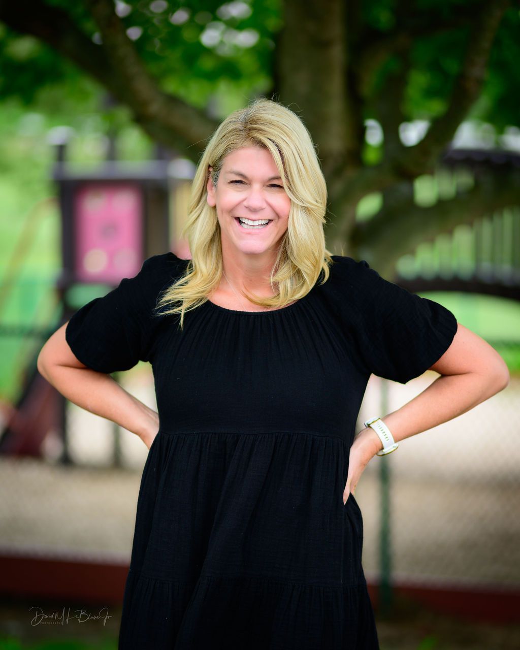 Blonde woman in black dress smiling, hands on hips, outdoors near a tree and a wooden structure.