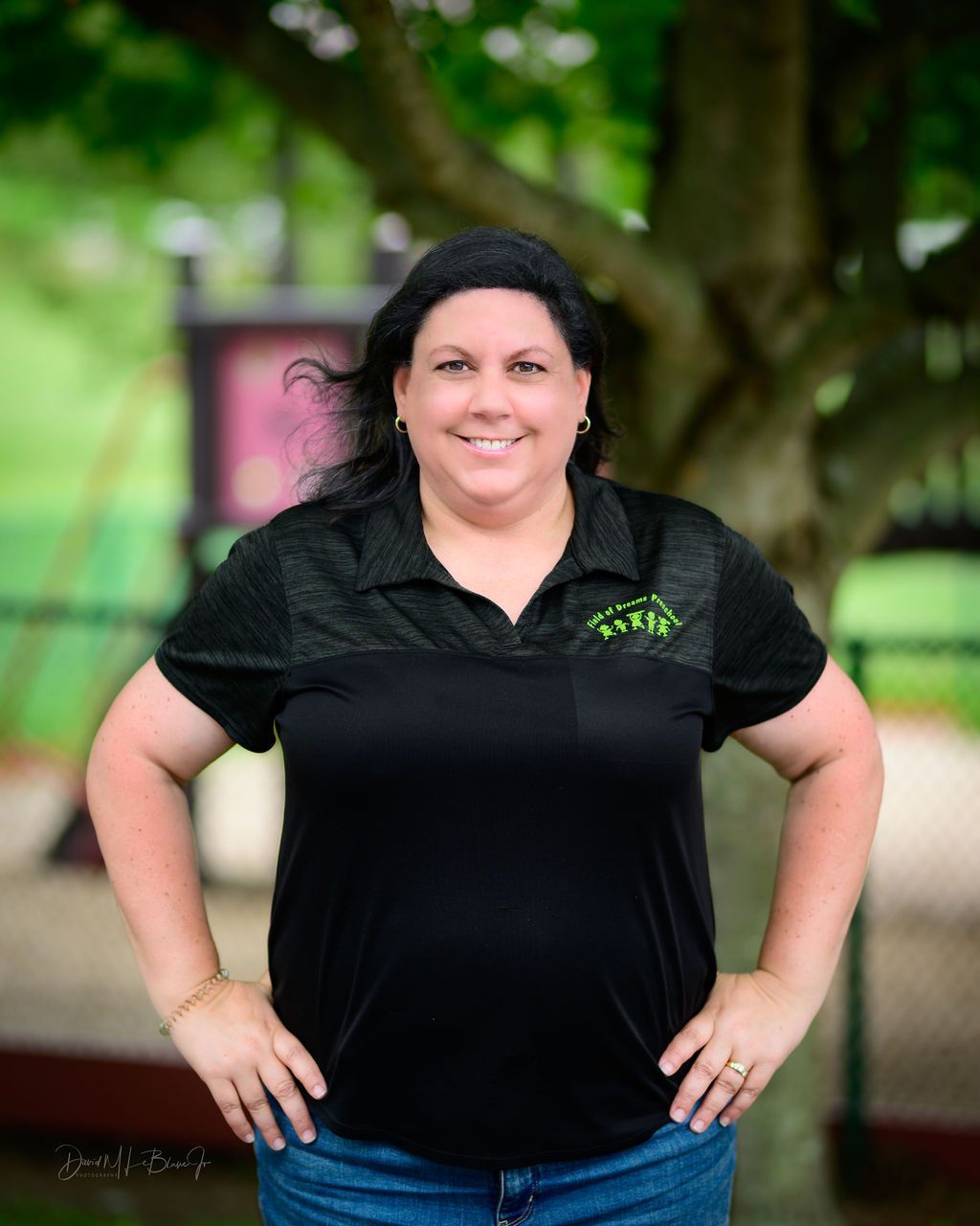 Woman with dark hair in black shirt, hands on hips, smiling outdoors near a tree.