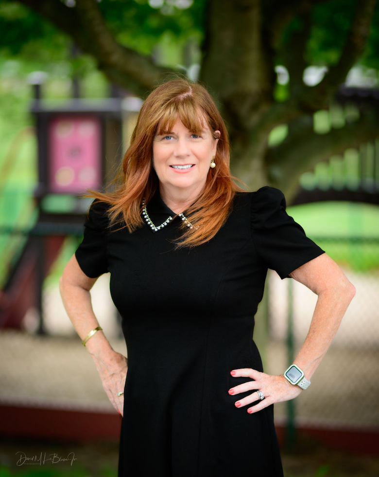 Woman with red hair in black dress, hands on hips, smiling outdoors.