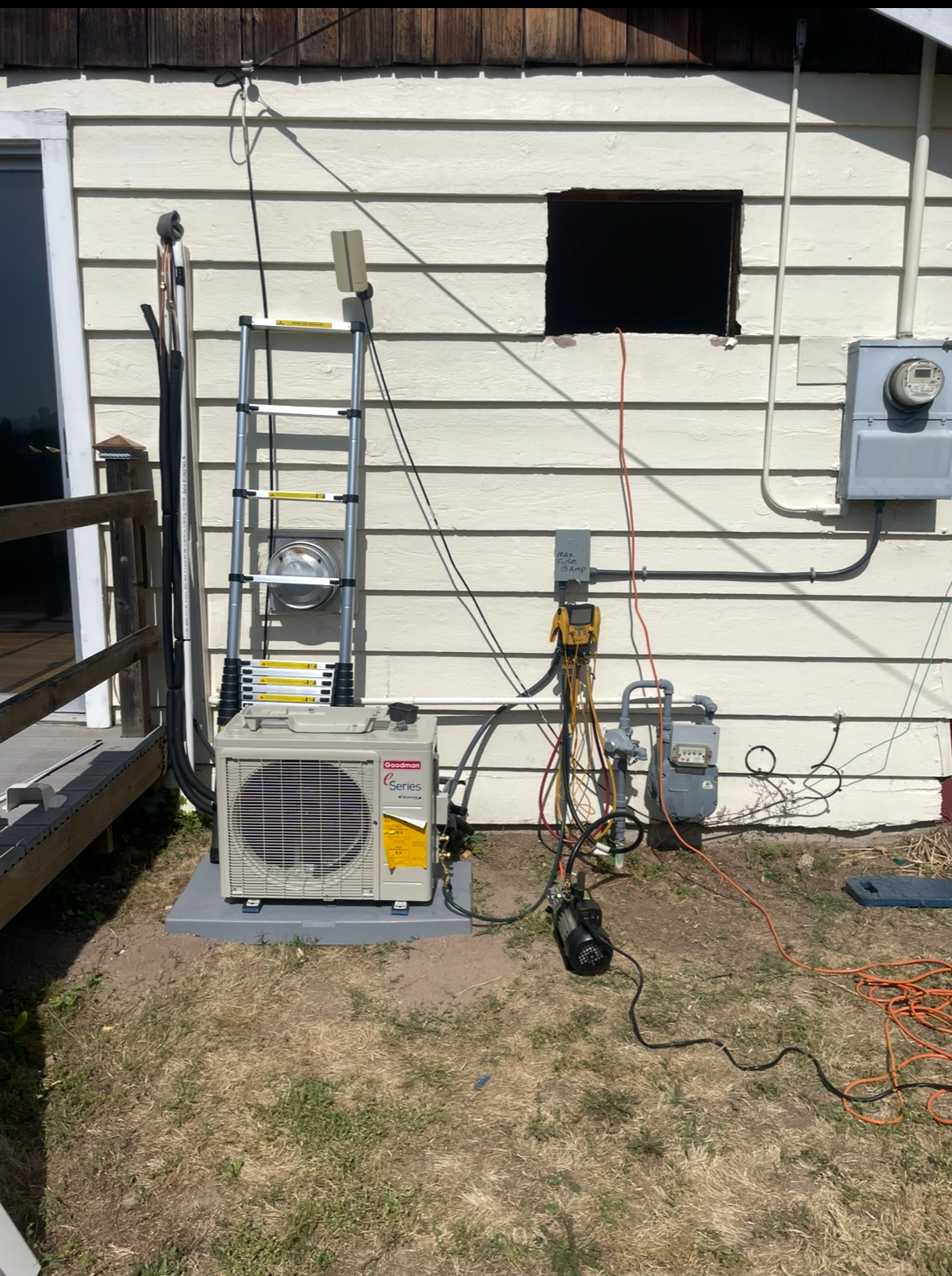 An AC unit with various wires and a ladder against a beige building, near a gas line and electrical box.