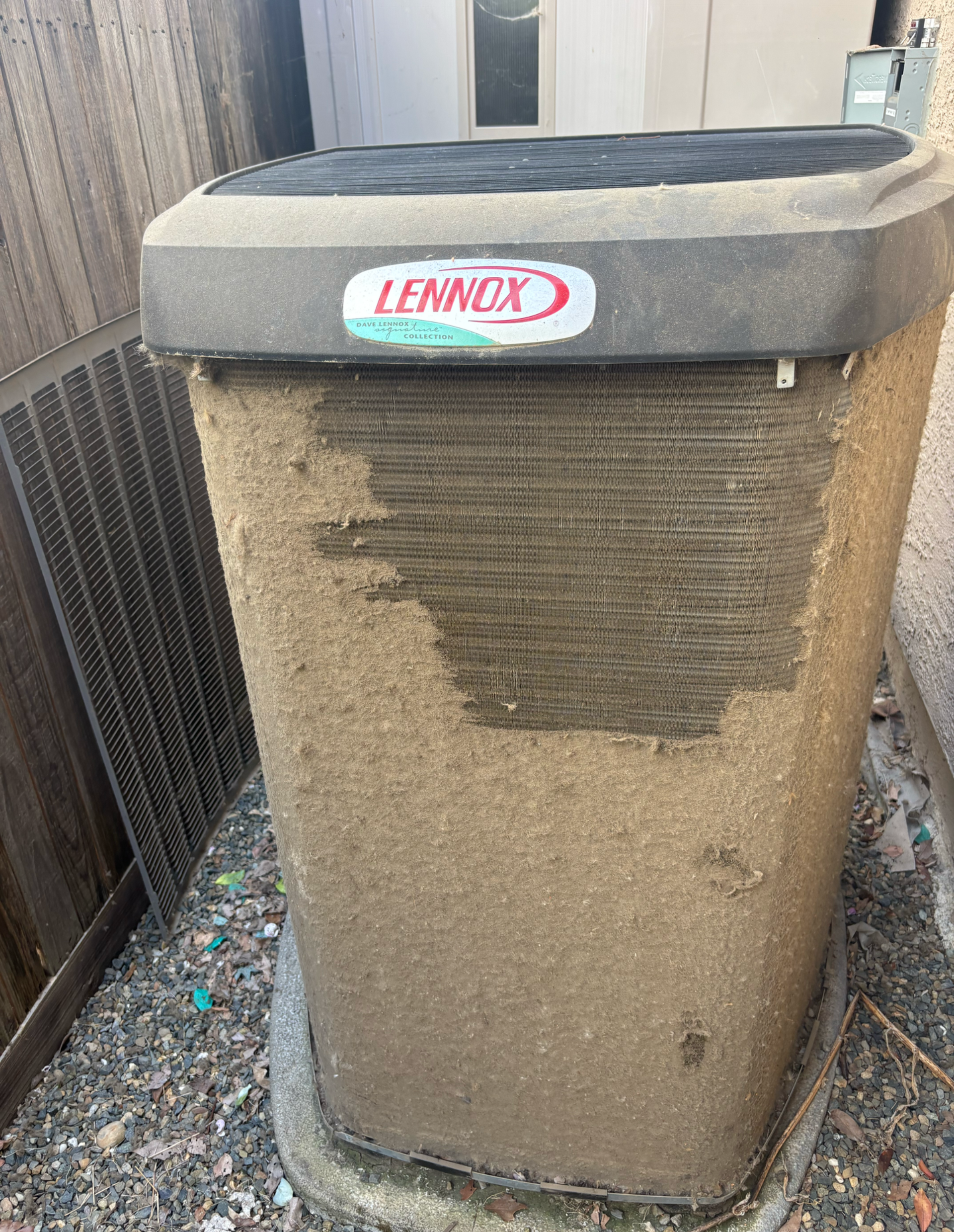 Dirty Lennox air conditioner unit, covered in dust, next to a building and grating.