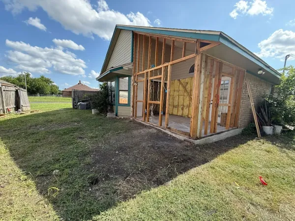 House undergoing renovation; exposed wooden frame, insulation visible, green yard. Blue sky and sun.