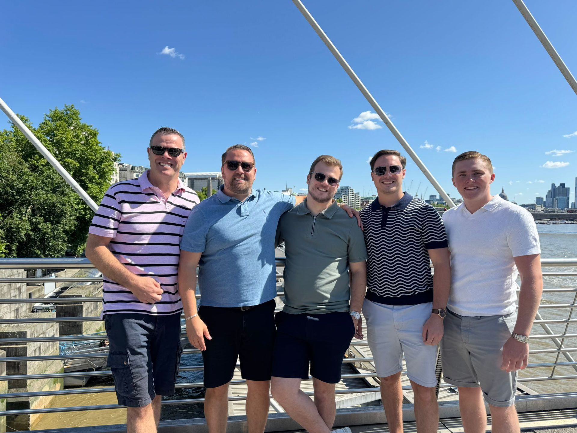 Five friends standing on a bridge, smiling in casual clothes on a sunny day.
