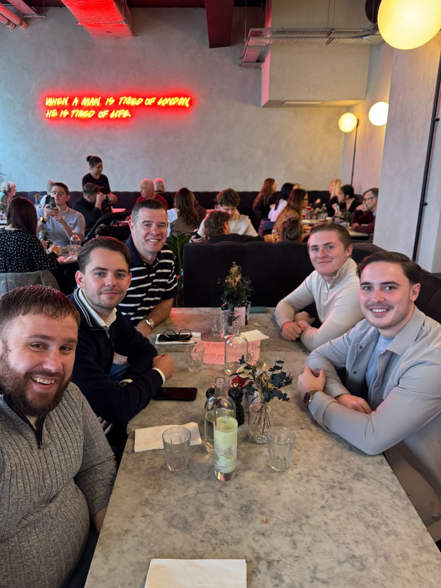 Group of friends smiling around a restaurant table with drinks in a cozy, busy dining room.