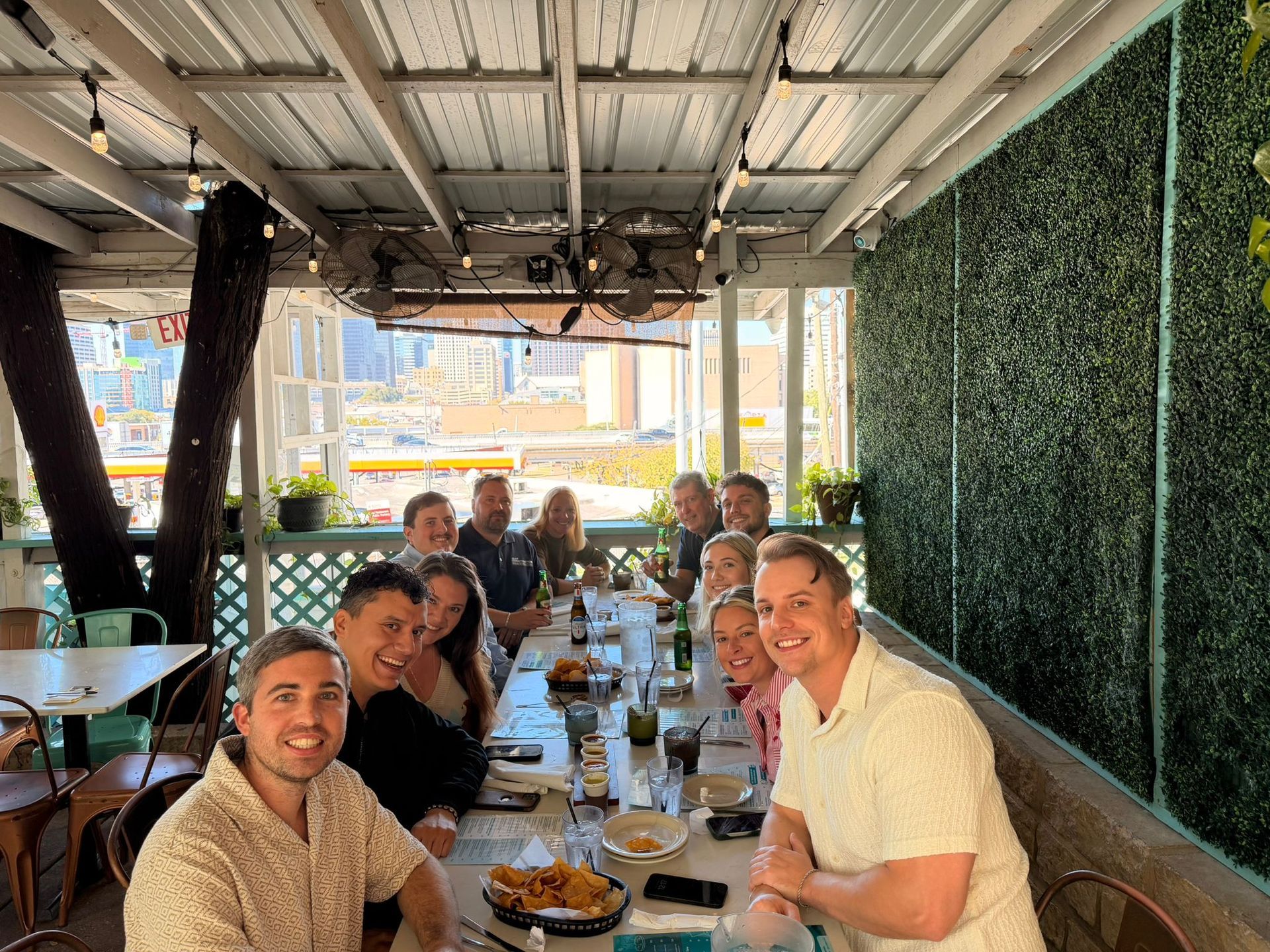 Group of friends sharing a meal at a long table in a bright outdoor restaurant, smiling at the camera