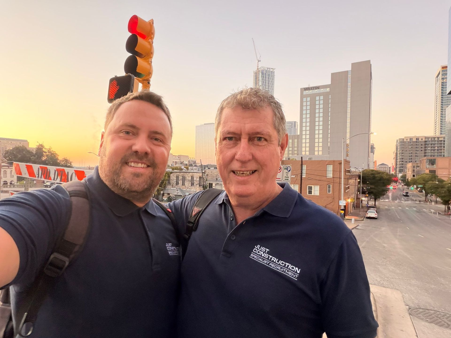 Two smiling men taking a selfie on a city street at sunset, with traffic lights and tall buildings behind them