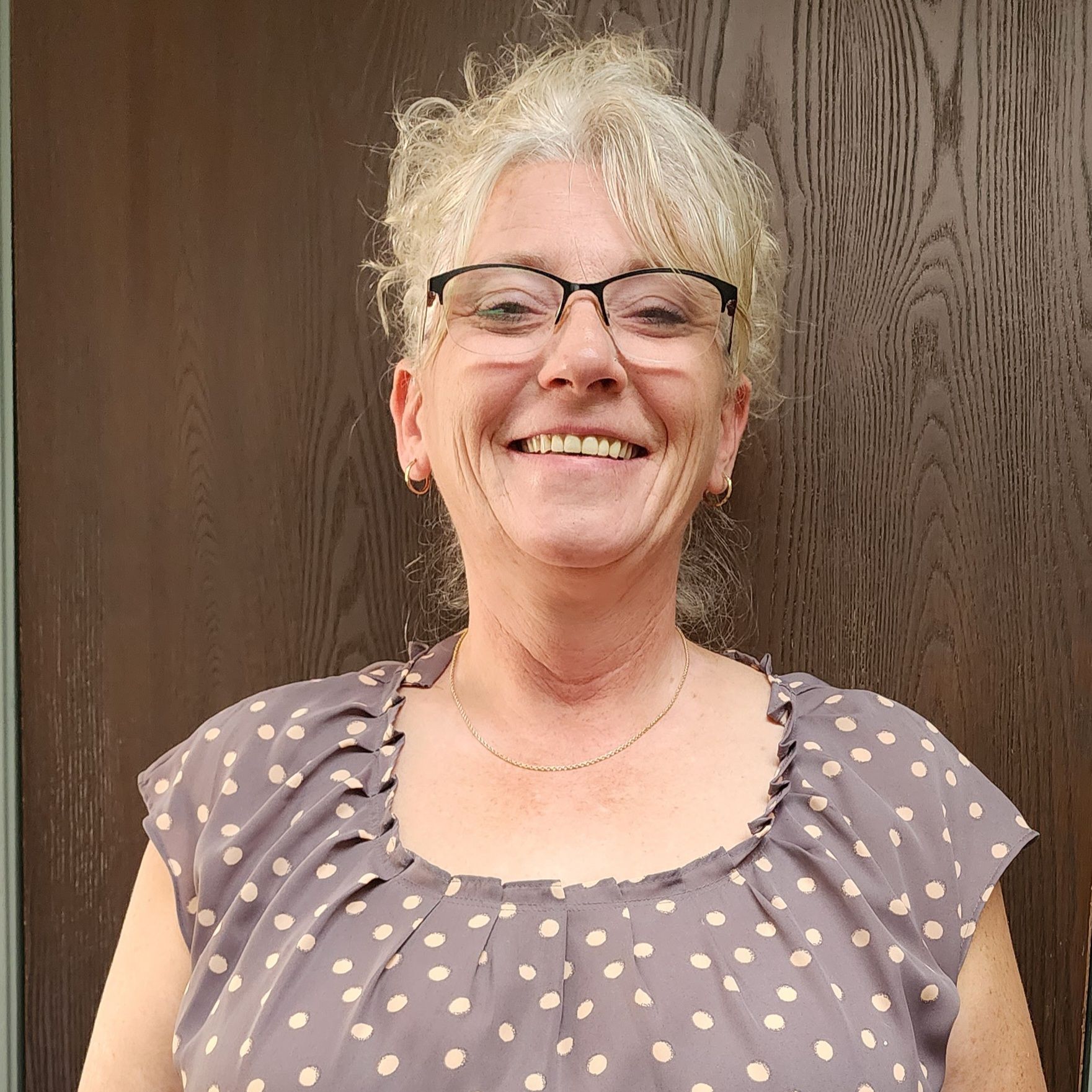 A woman wearing glasses and a polka dot shirt is smiling in front of a wooden door.