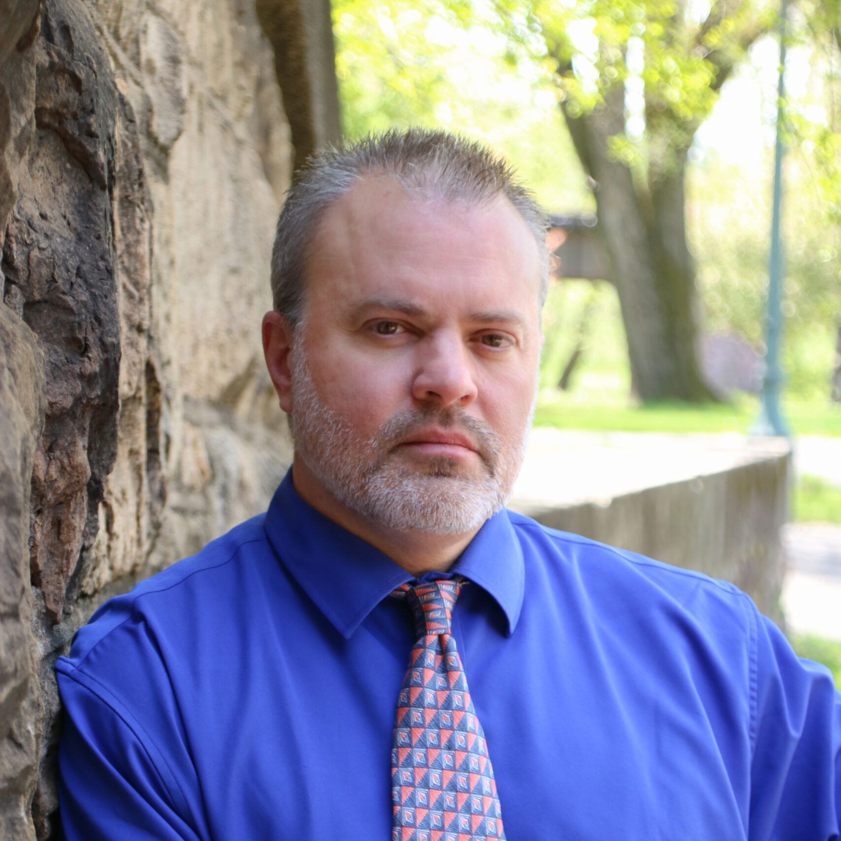 A man in a blue shirt and tie leans against a stone wall