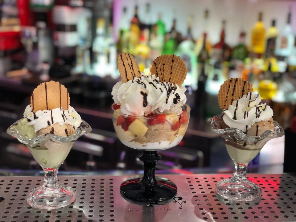 Tre coppe gelato con panna montata, frutta e biscotti waffle su un bancone.