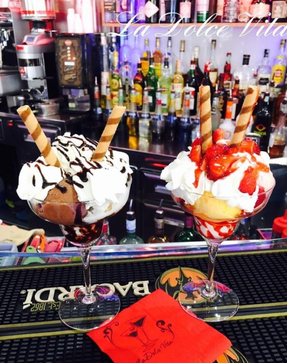 Due coppe gelato con panna montata, cialde e guarnizioni, su un bancone da bar con bottiglie.