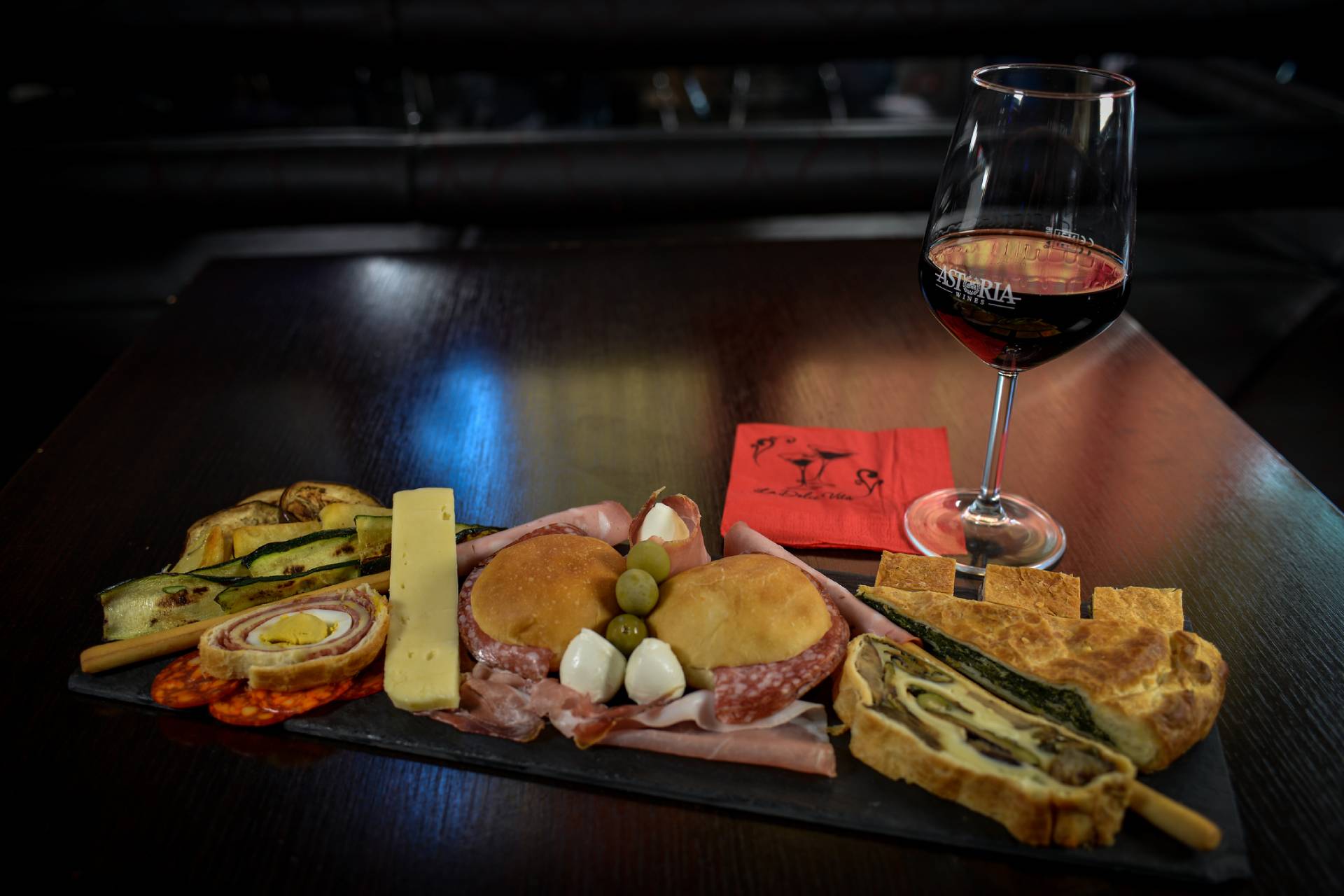 Un tagliere di salumi con vino e vari cibi italiani su un tavolo buio.