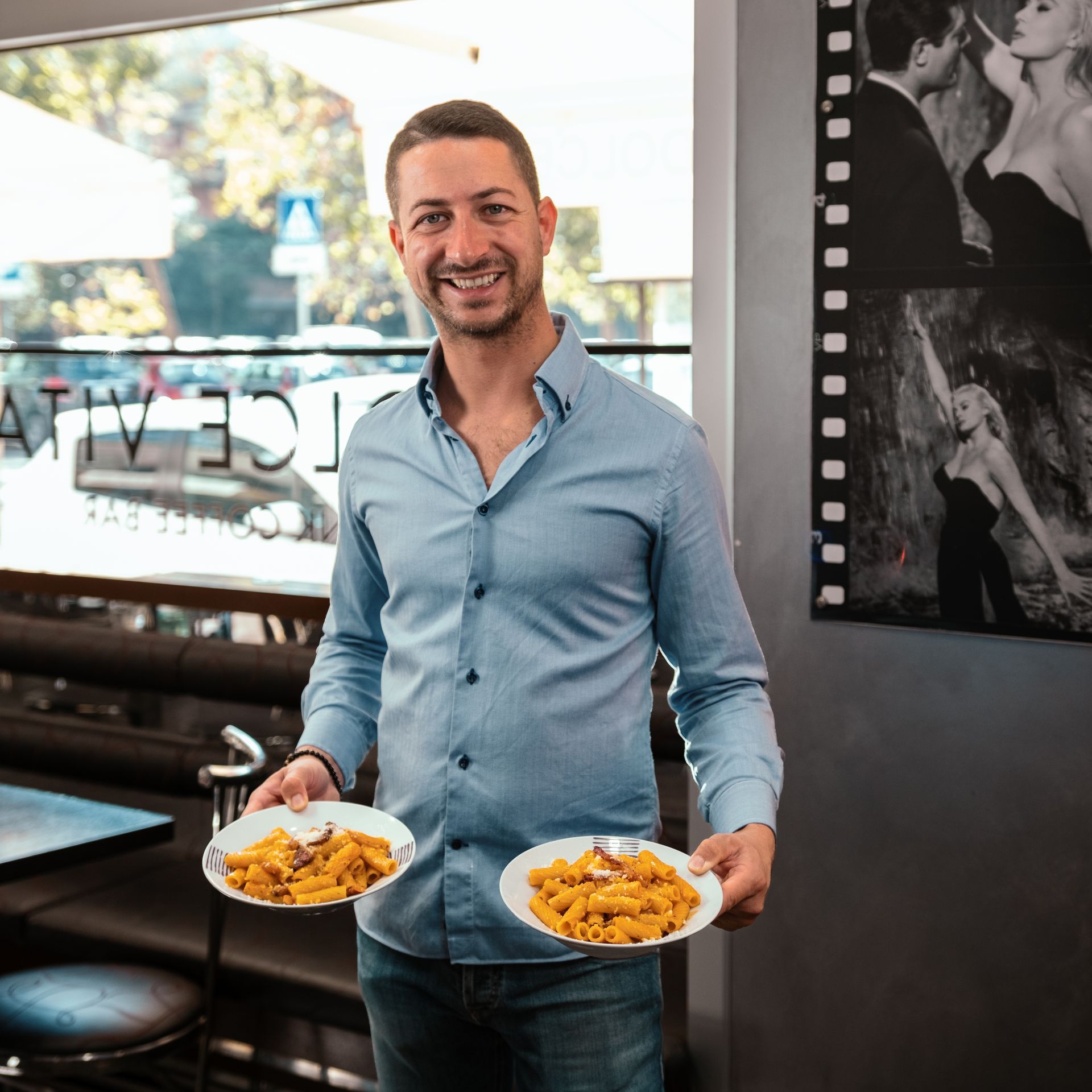 Un uomo con una camicia blu sorride, tenendo in mano due piatti di pasta in un ristorante.