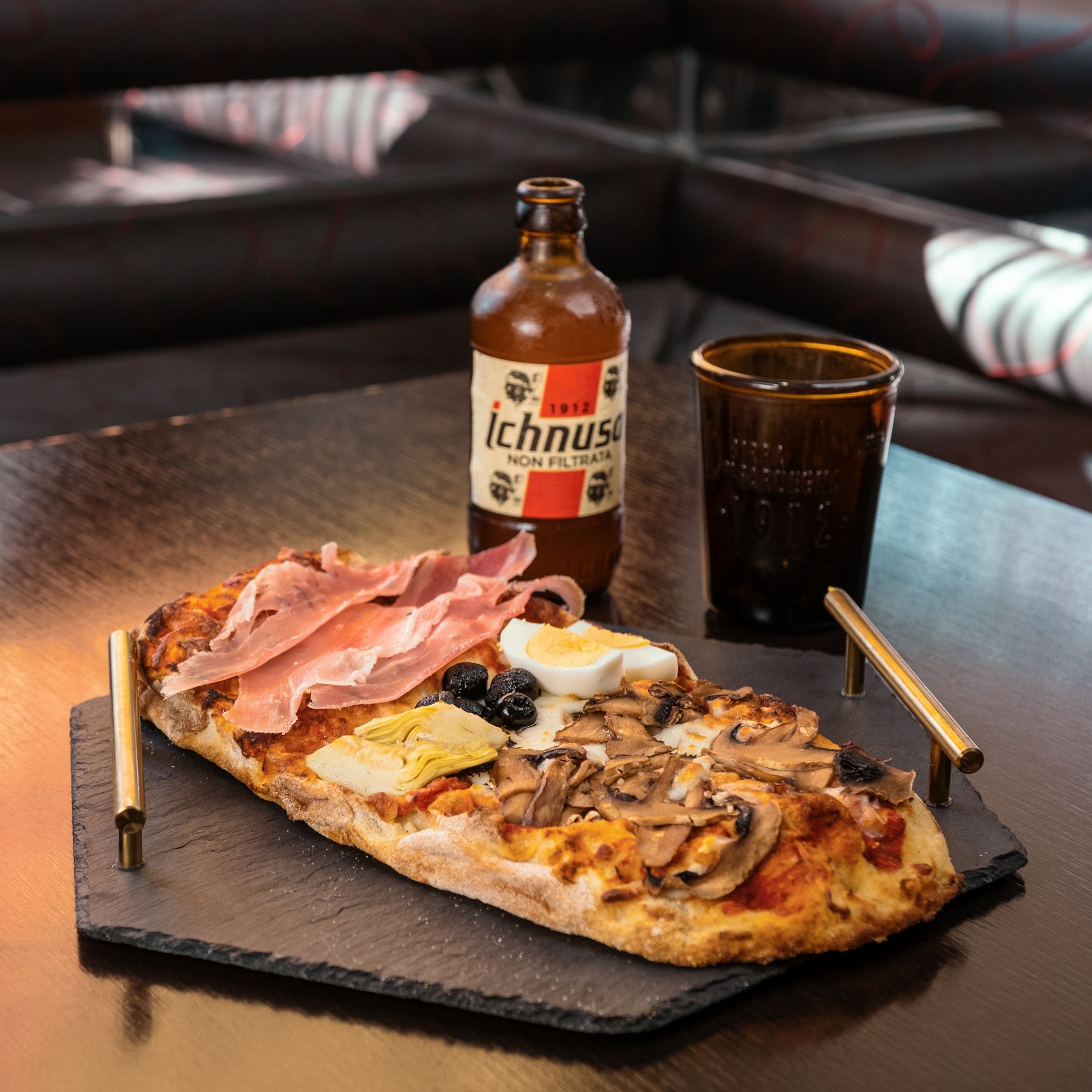 Pizza con condimenti, bottiglia di birra e bicchiere su un vassoio. Ambiente buio.