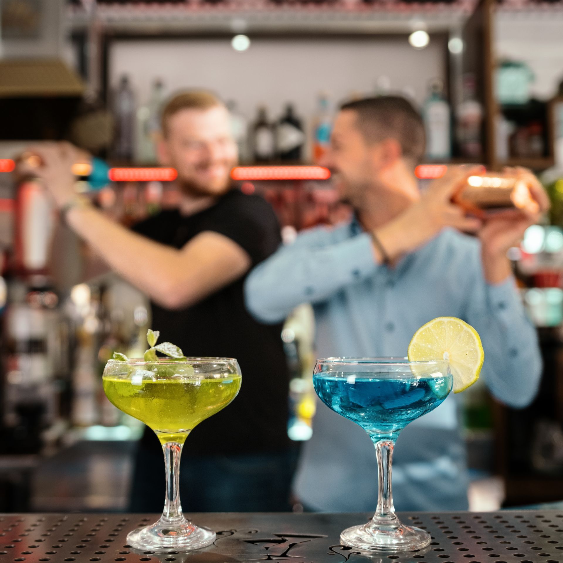 Due cocktail sul bancone di un bar, con i baristi che shakerano i drink sullo sfondo. Drink verdi e blu.