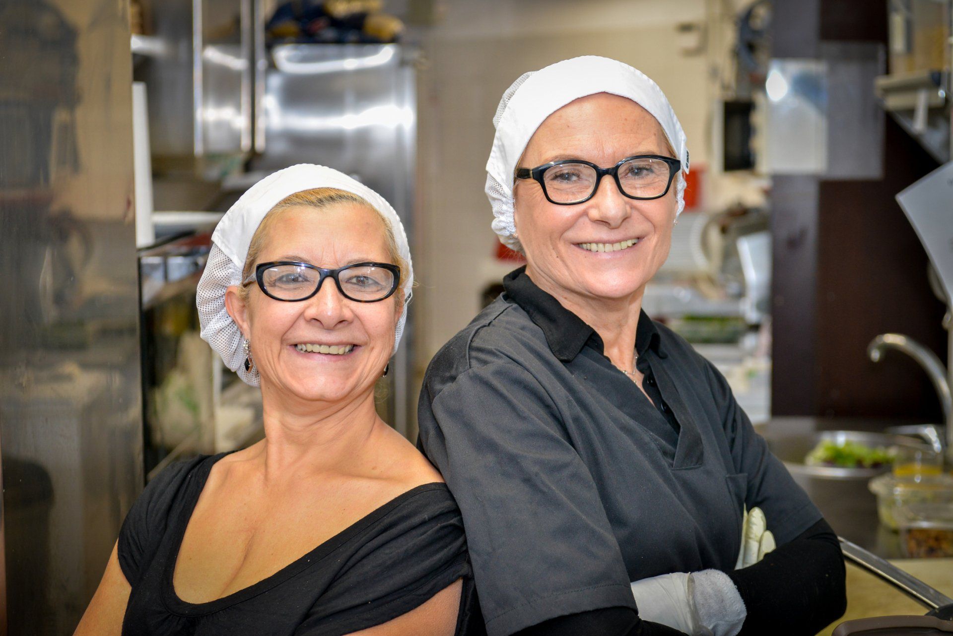 Due persone con retine per capelli e occhiali sorridono in una cucina commerciale.