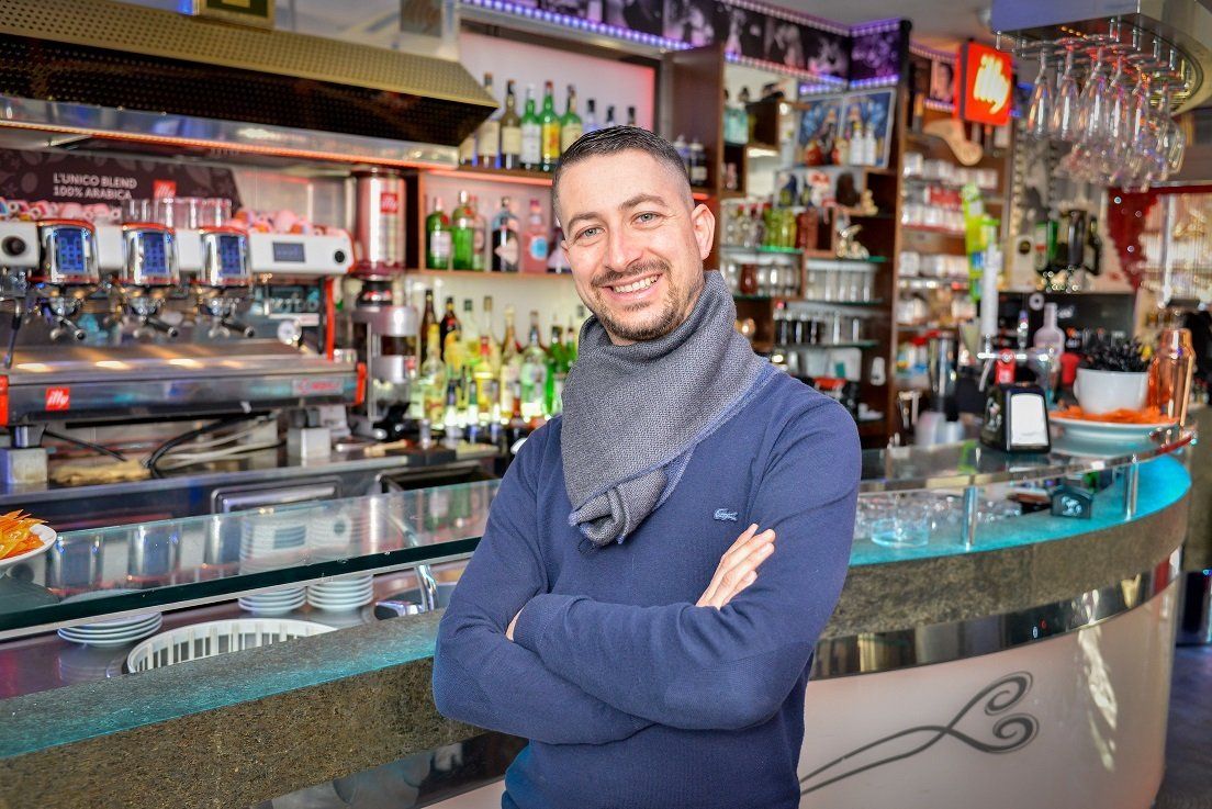 Uomo sorridente con le braccia incrociate, in piedi davanti a un bar con bottiglie e una macchina per il caffè.