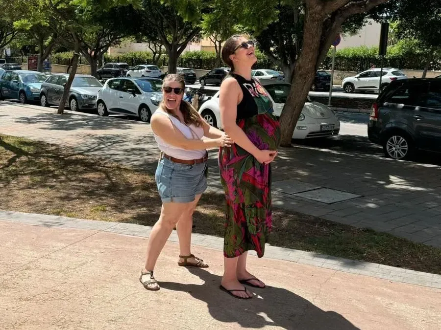 Dos mujeres de pie en una acera soleada, una sujetando a la otra por detrás, con coches aparcados y árboles cerca.