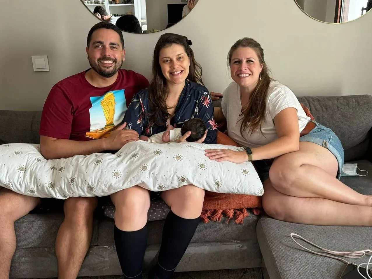 Tres amigas sonriendo en un sofá, sosteniendo un perro blanco y posando en una sala de estar.