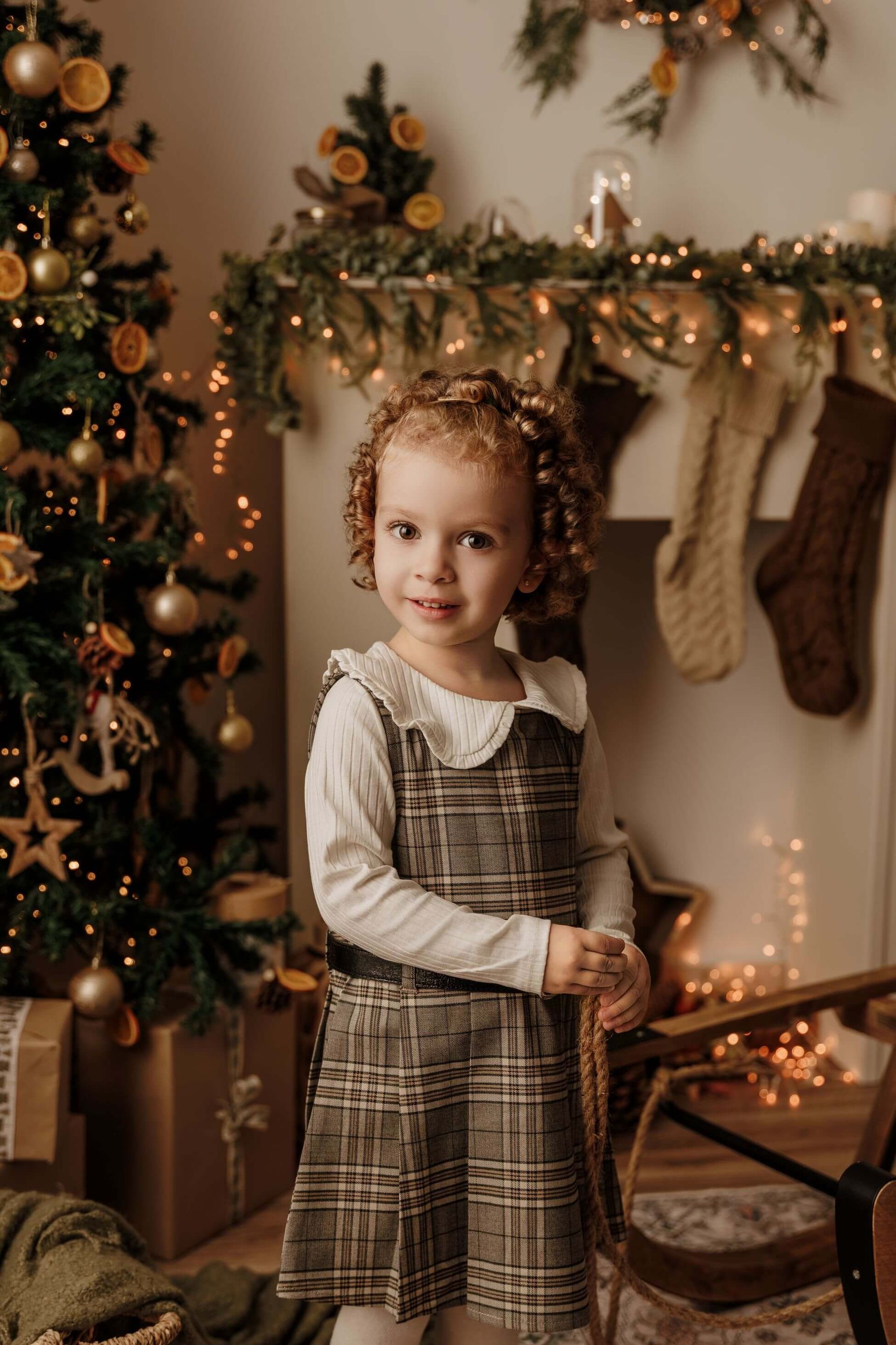Un niño de pie junto a un árbol de Navidad decorado y una chimenea en una acogedora habitación navideña con una cálida iluminación.
