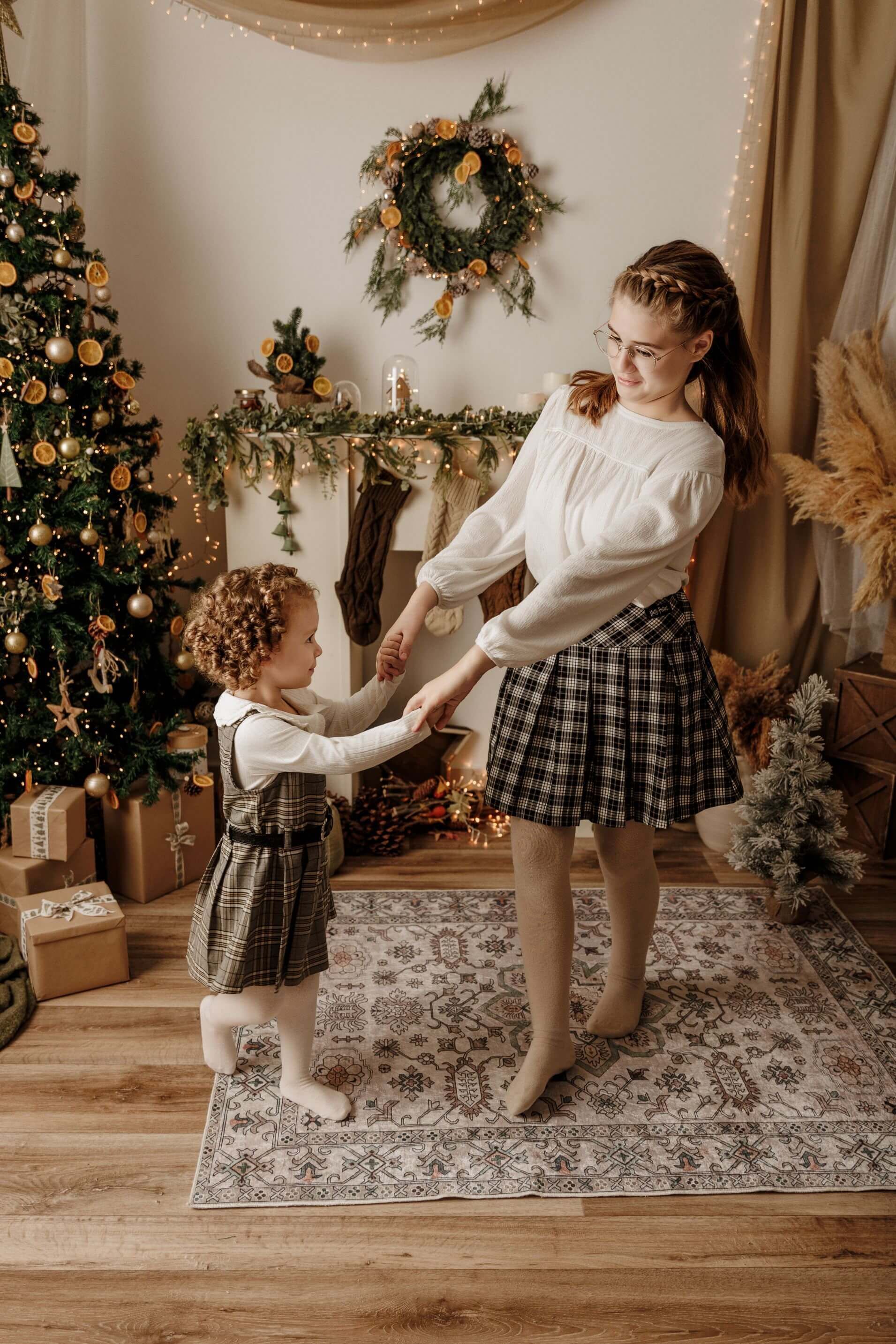 Dos niños tomados de la mano en una sala festiva decorada con un árbol de Navidad, una corona y regalos envueltos.