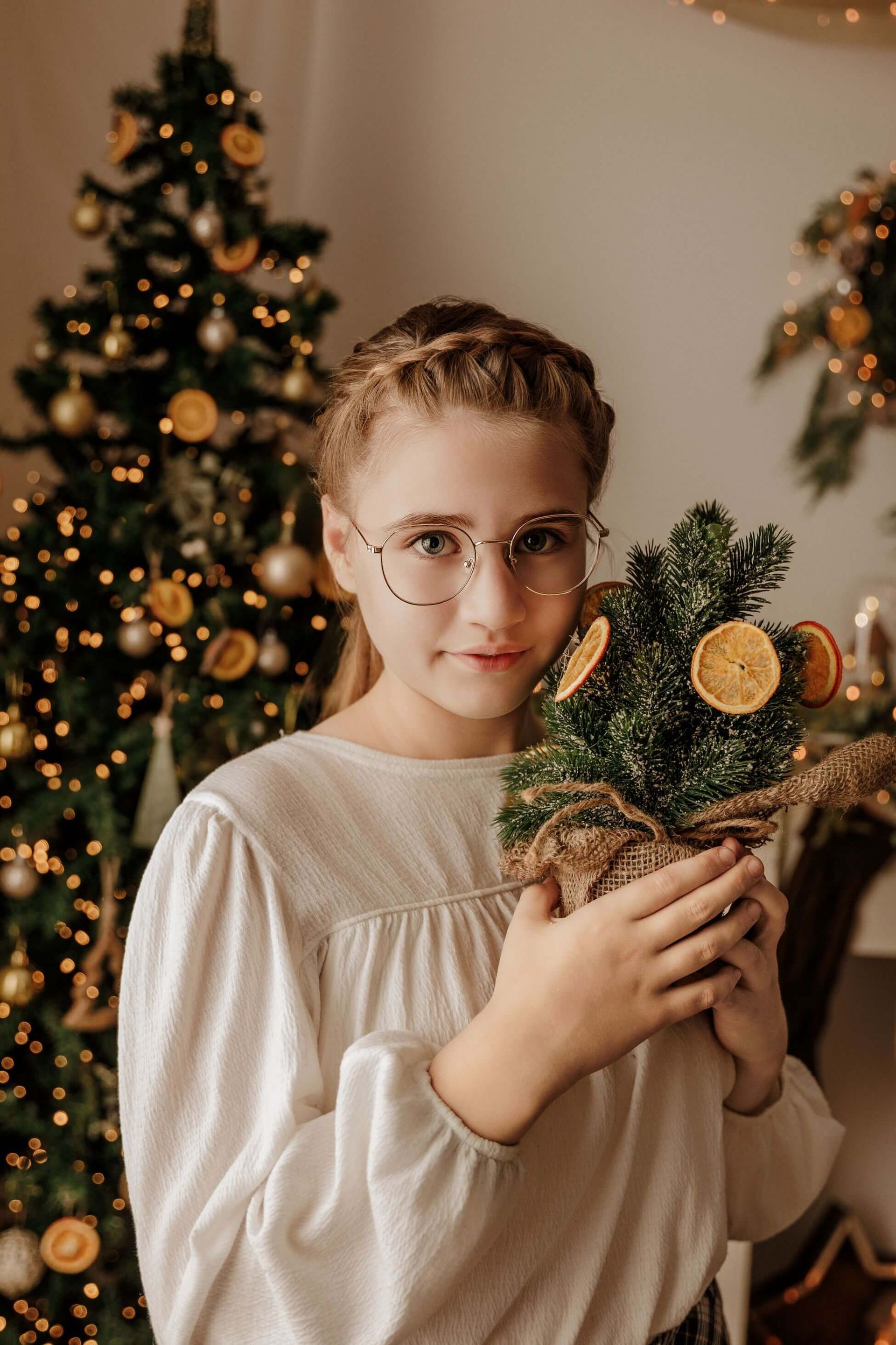 Una persona sostiene un pequeño arreglo floral de pino decorado frente a un árbol de Navidad iluminado.