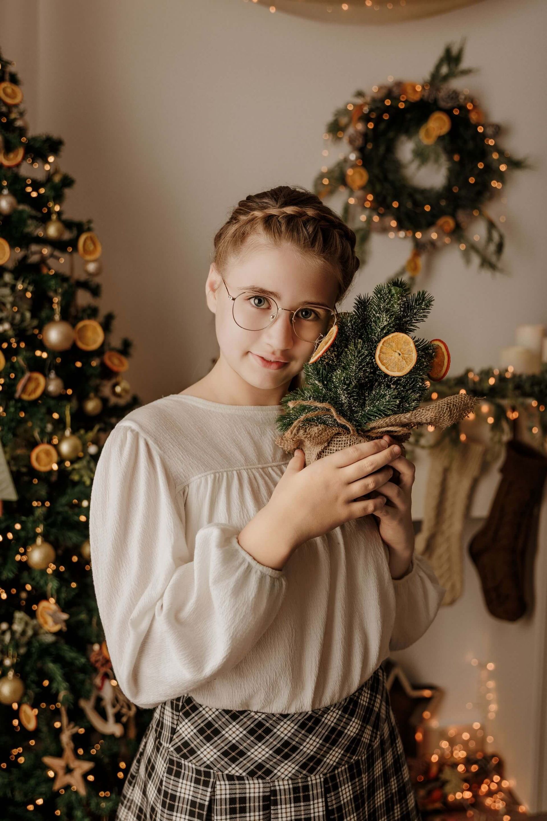 Una persona sostiene un pequeño árbol de Navidad decorado junto a una corona navideña iluminada y un árbol en el interior de una casa.