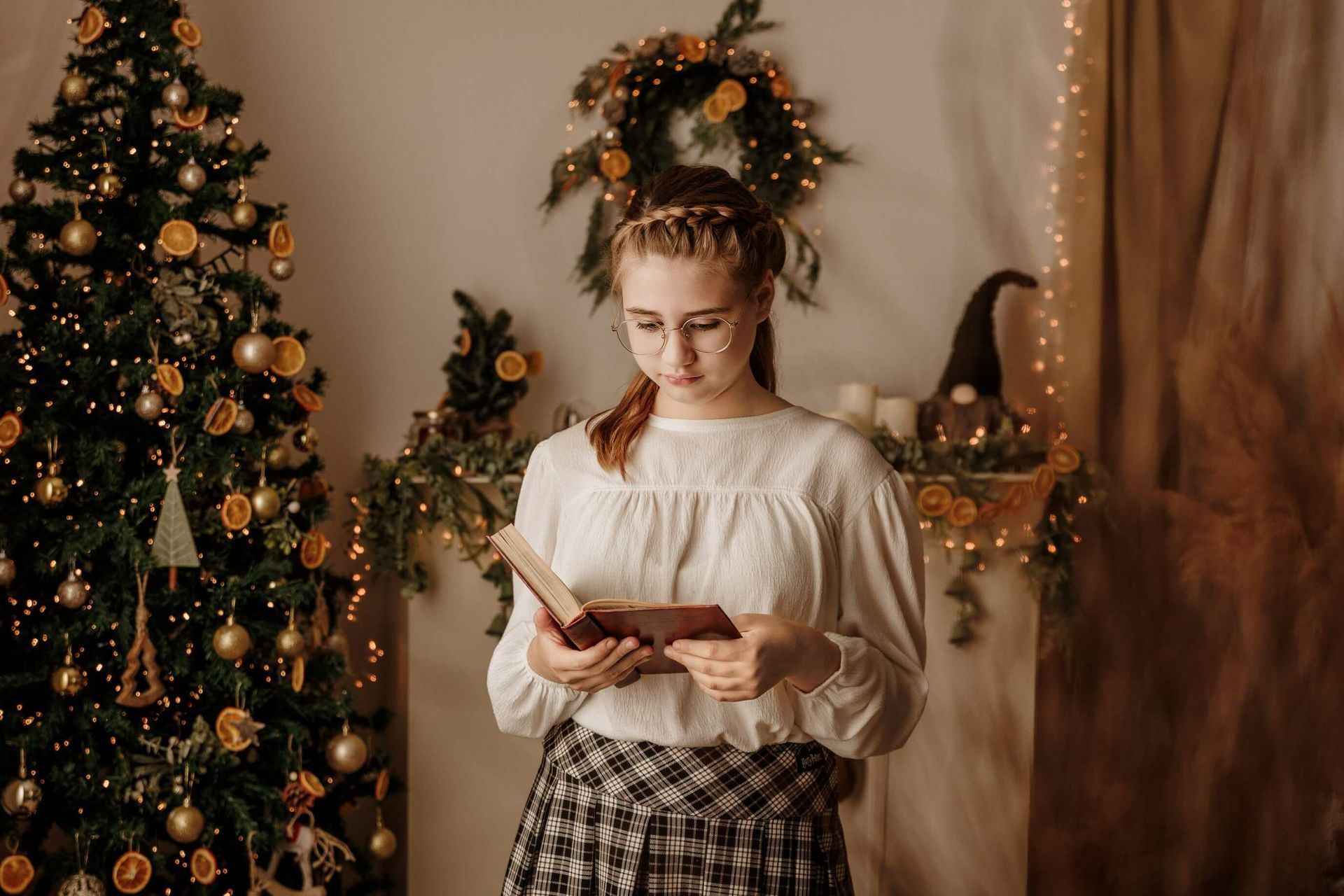Mujer leyendo un libro junto a un árbol de Navidad decorado y coronas navideñas.