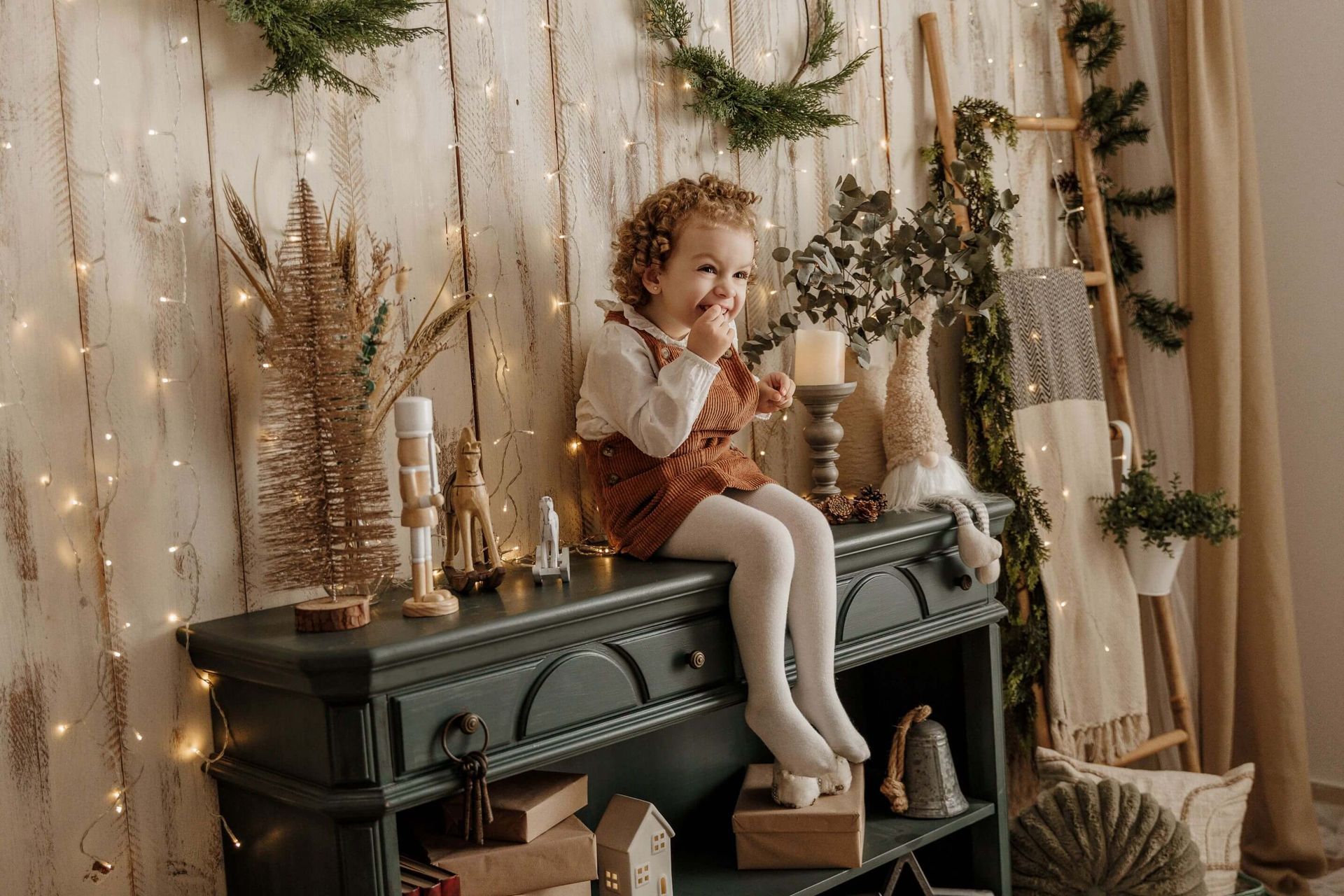 Un niño sentado sobre un armario verde decorado en una habitación festiva con velas, plantas secas y luces tenues.