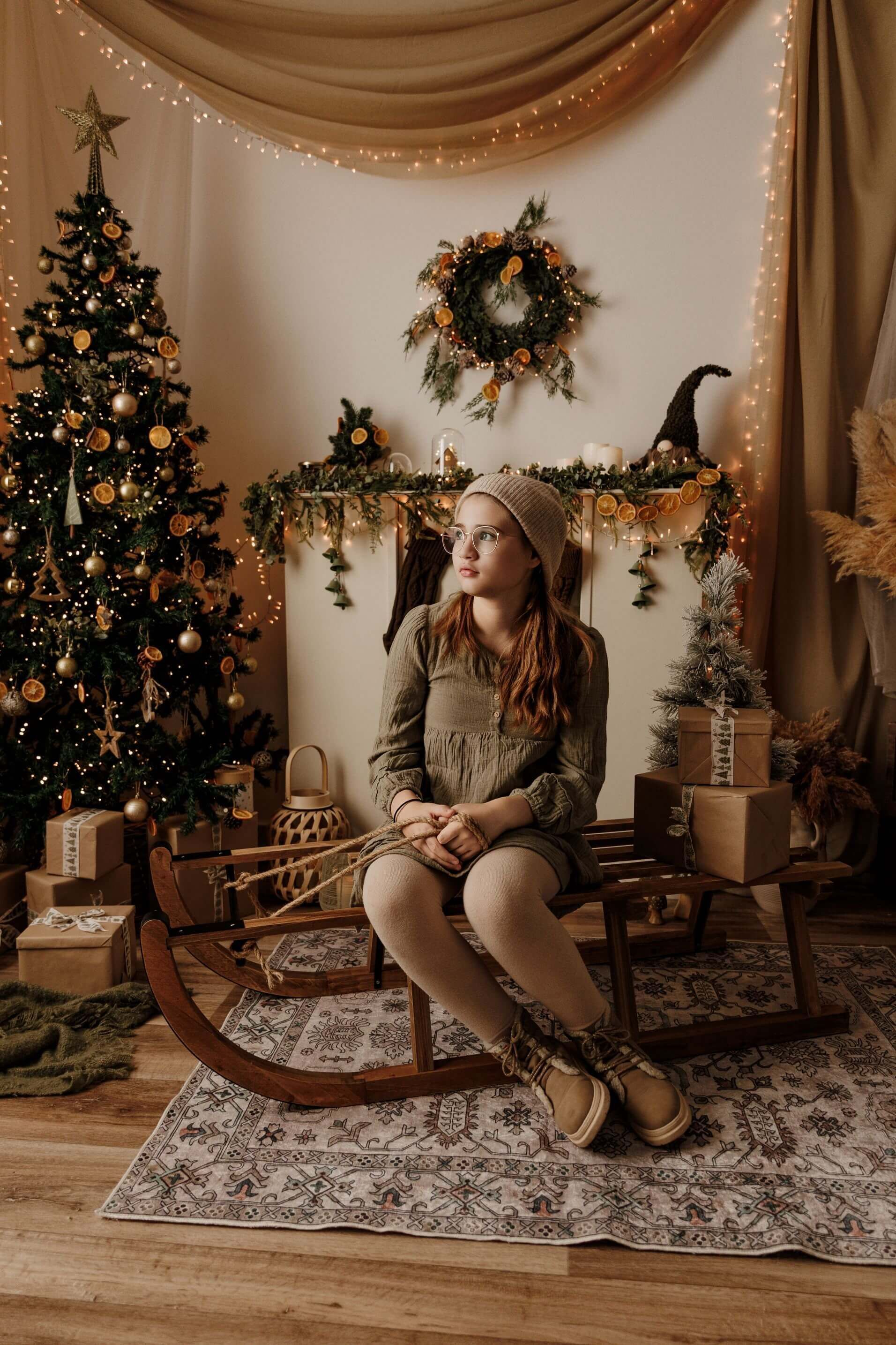 Niña sentada junto a un árbol de Navidad decorado y una chimenea, con un vestido festivo en una acogedora habitación navideña.