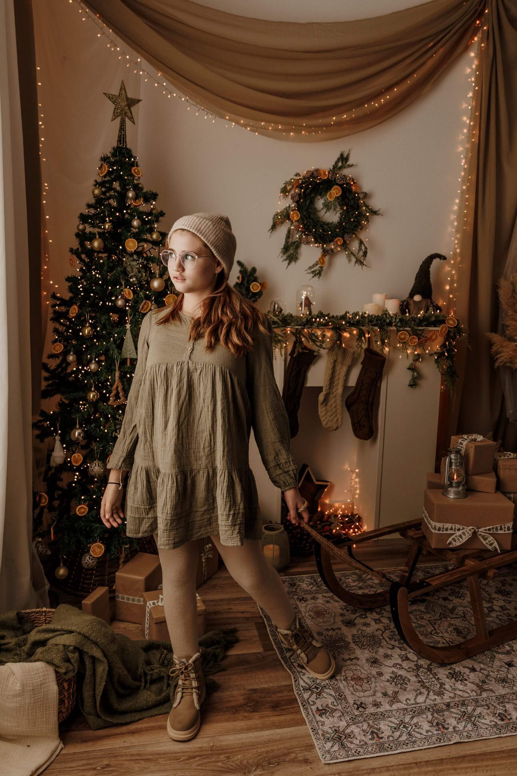 Mujer de pie en una acogedora habitación decorada para Navidad, junto a un árbol iluminado y una repisa de chimenea con calcetines navideños.