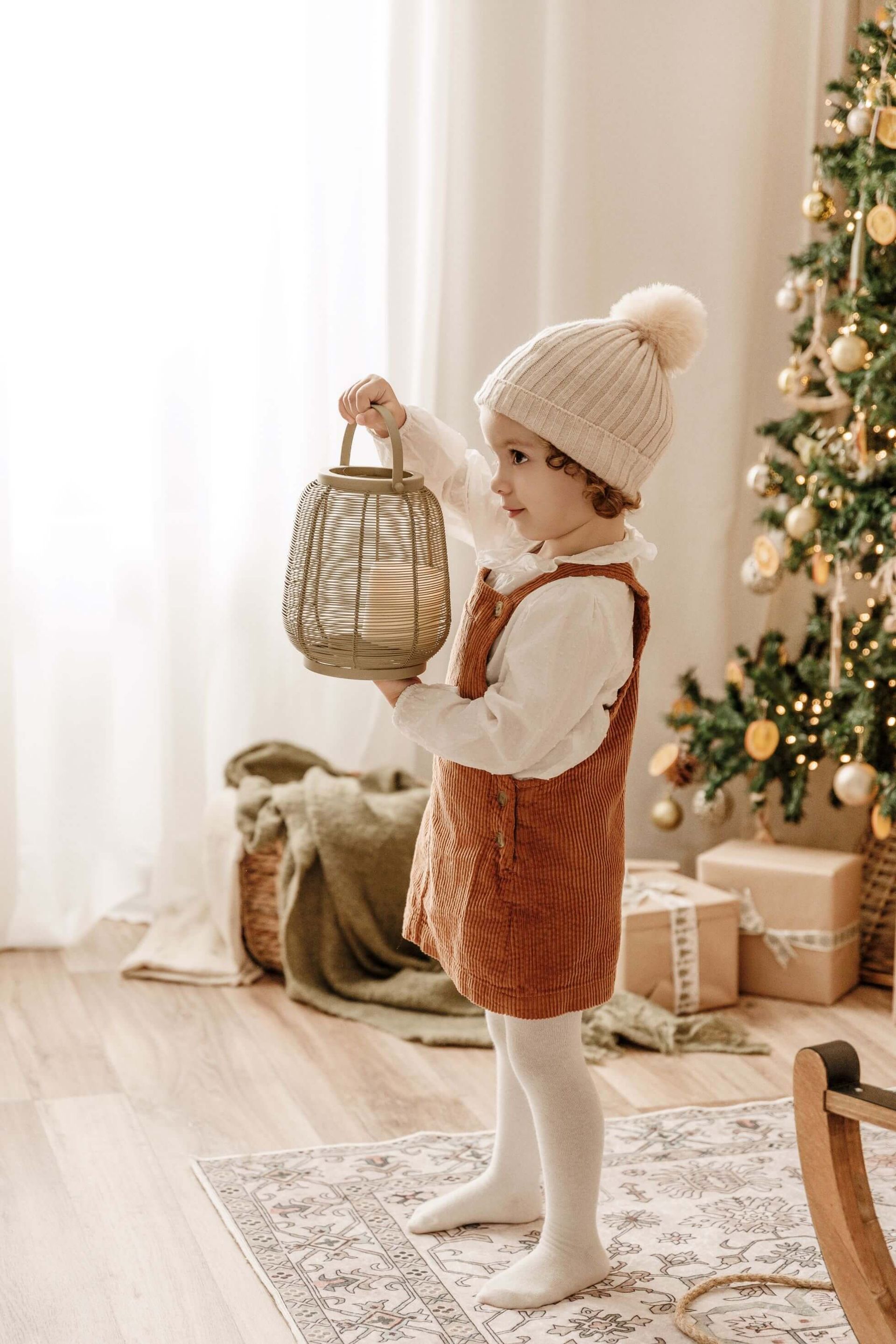 Un niño sostiene una linterna de mimbre junto a un árbol de Navidad decorado en una habitación acogedora.