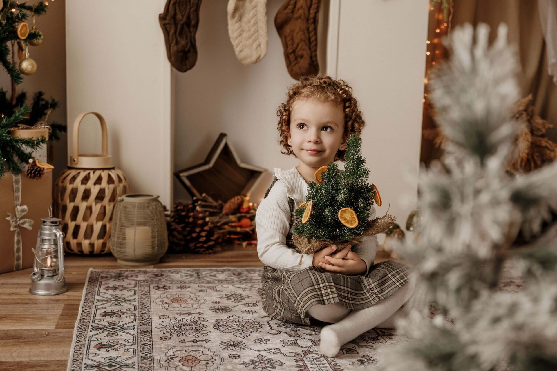 Un niño sostiene un pequeño árbol de Navidad junto a una chimenea decorada con luces cálidas y una alfombra estampada.