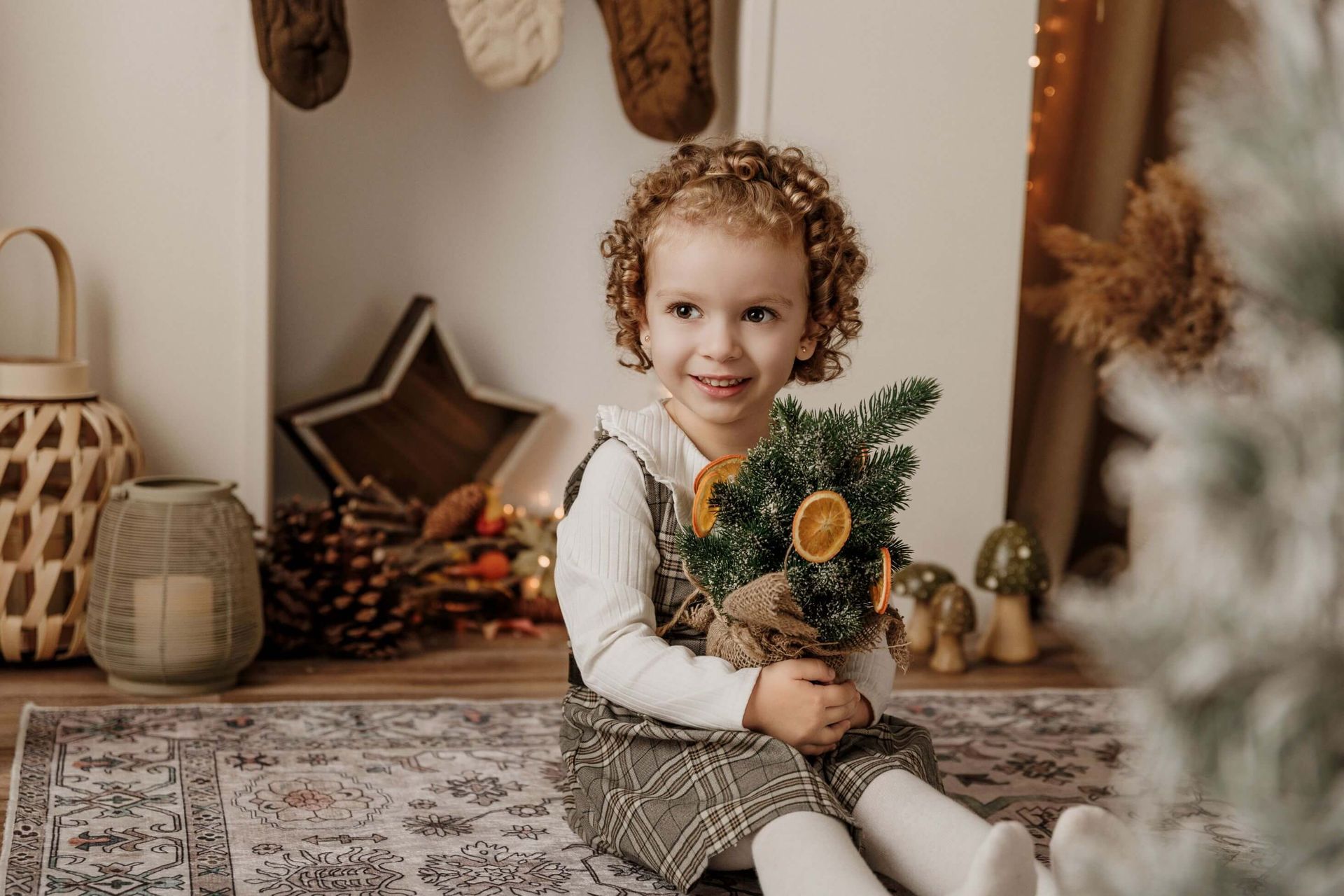 Un niño pequeño sostiene un pequeño árbol de Navidad en una acogedora habitación navideña, sonriendo sobre una alfombra estampada.