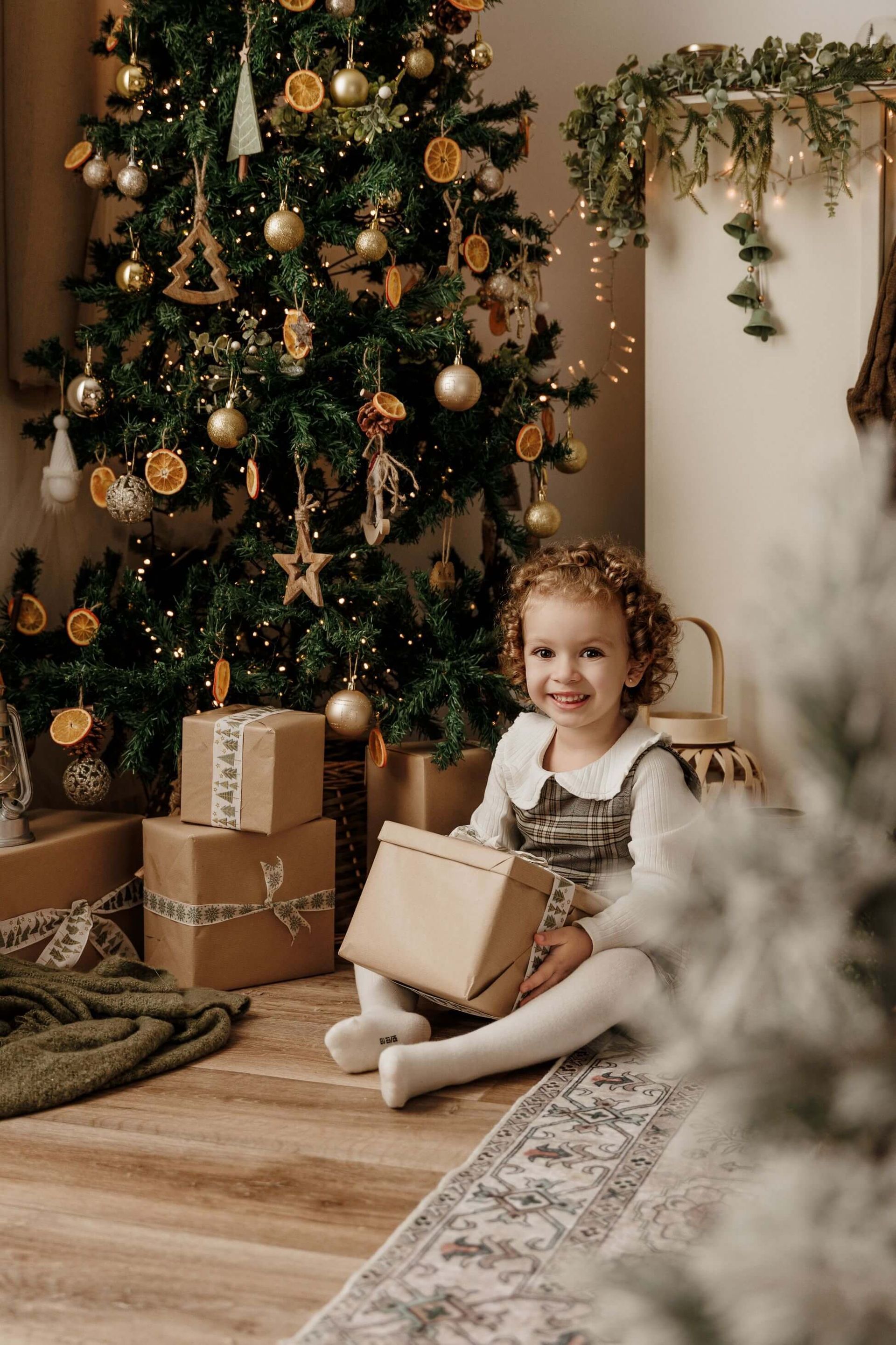 Un niño sentado junto a un árbol de Navidad decorado con regalos envueltos, sonriendo en una acogedora sala de estar.