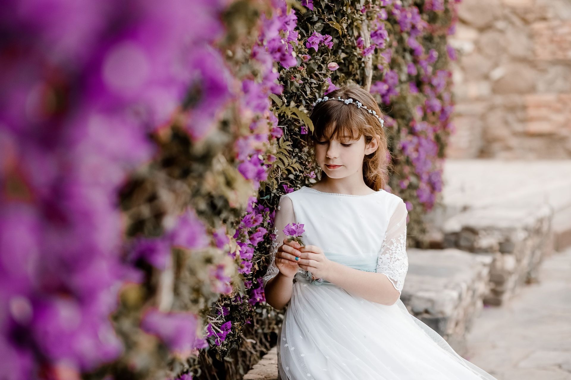 Una persona vestida de blanco sostiene flores junto a una pared cubierta de flores moradas.